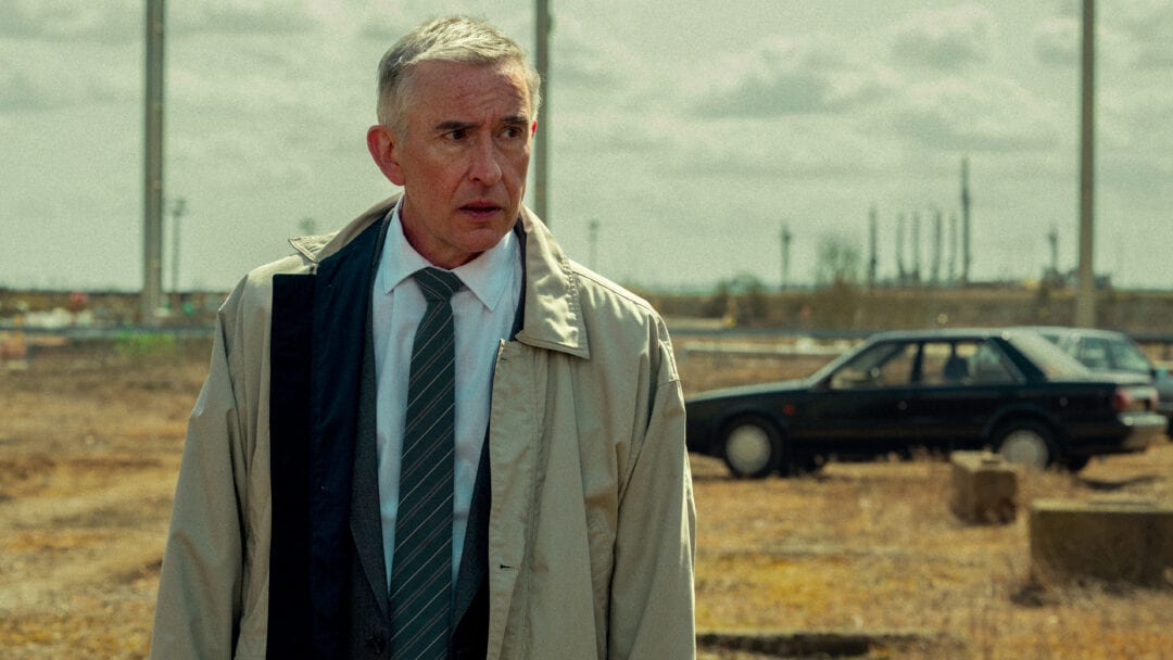 "Middle-aged man in a trench coat and tie stands in an empty, industrial field with a dark car and refinery structures in the background, under a cloudy sky."
